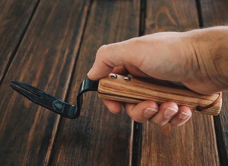 May include: A black metal hand weeding tool with a wooden handle. The handle is made of light and dark wood with a leather strap.