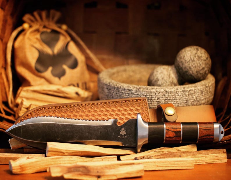 May include: A black and silver knife with a wooden handle, resting on a pile of wooden sticks. The knife has a serrated blade and a leather sheath. The knife is surrounded by other items, including a small bag and a bowl.