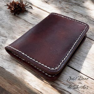 May include: A dark brown leather wallet with white stitching. The rectangular wallet is closed. The text "Just Beer & Skittles" is visible. The wallet is on a wooden surface. The wallet is likely to be approximately 10 cm x 8 cm.
