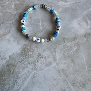 May include: A beaded bracelet featuring a mix of blue, white, and clear beads. The bracelet includes beads with heart and cross designs. The beads are strung together with a clear elastic cord. The bracelet is displayed on a marble surface.