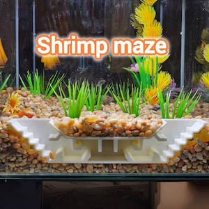 May include: A white plastic shrimp maze with steps and a rectangular base is set in a fish tank. The tank contains small, light brown pebbles, green plastic plants, and yellow and pink artificial plants. The text "Shrimp maze" is displayed in orange.