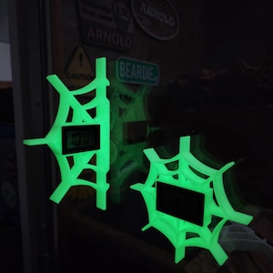 May include: Two glowing green spiderweb-shaped digital clocks. The clocks display the time and are mounted on a dark surface. The spiderweb design is intricate and detailed, with a bright, neon-green color.