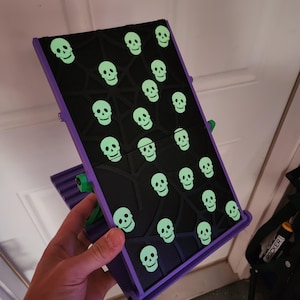 May include: A purple and black tablet stand featuring a pattern of glowing green skulls. The stand has a textured black surface with a geometric design, and the skulls are evenly spaced across the surface. The stand is held open by a green hinge.