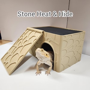 May include: Beige reptile habitat with a textured stone-like exterior and a black top. A bearded dragon is inside the habitat, with its head and front legs visible. The text "Stone Heat & Hide" is above the habitat.