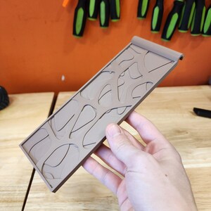 May include: A tan-colored, rectangular tray with a unique, organic cutout design. The tray has a raised edge on one side and is held by a person's hand. The background includes tools and a wooden surface.