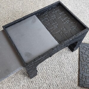 Może przedstawiać: Czarny drewniany stołek z odpinanym siedziskiem. Stołek ma fakturowaną powierzchnię przypominającą kamień. Siedzisko jest szare i ma gładką powierzchnię.