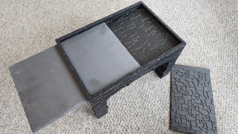 Może przedstawiać: Czarny drewniany stołek z odpinanym siedziskiem. Stołek ma fakturowaną powierzchnię przypominającą kamień. Siedzisko jest szare i ma gładką powierzchnię.