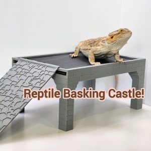 May include: A gray reptile basking castle with a ramp. The castle has a flat top and a brick-like design. A bearded dragon lizard is sitting on the top of the castle. The text "Reptile Basking Castle!" is written in brown letters on the front of the castle.