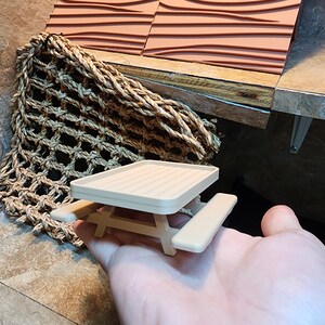 May include: A miniature beige wooden picnic table with a rectangular top and two benches. The table is small enough to fit in the palm of a hand.