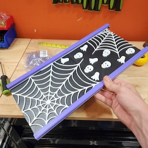 May include: A Halloween-themed decorative tray with a black and white spiderweb design. The tray features white ghost and skull accents, with a purple border. The tray is designed for Halloween decorations.
