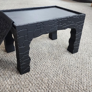 Może przedstawiać: Czarny, składany stolik z fakturowaną powierzchnią. Stolik ma płaski blat i cztery nogi.
