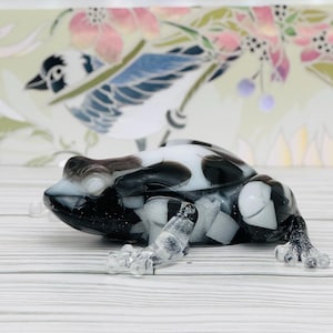 May include: A decorative glass frog figurine in black and white. The frog has a speckled pattern on its back and clear legs. The background features a floral and bird design in soft colors.