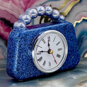 May include: A blue glitter handbag-shaped clock with a silver-tone clock face. The clock face has black Roman numerals and black hands, with a red second hand. The handle is made of silver-colored, spherical beads.