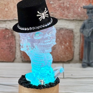 May include: A glowing blue skeleton figurine wearing a black top hat with a silver spider embellishment. The figurine is sitting on a wooden base with black rocks and a silver spider.