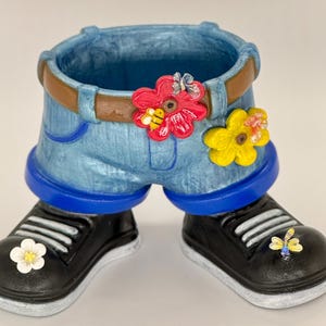 May include: A whimsical planter shaped like denim shorts with a brown belt, blue trim, and black sneakers. The shorts are adorned with red and yellow flower accents, and the sneakers have flower and butterfly details. A fun, decorative item.