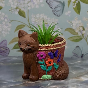 May include: A brown fox-shaped planter with a woven texture. The planter features a decorative design of flowers and butterflies in shades of purple, orange, and blue. It contains a green plant. The planter is a decorative item for home decor.