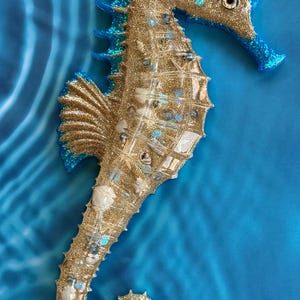 May include: A seahorse wall decoration featuring a gold glitter body with blue glitter accents along the fins and head. The surface is embedded with various small white seashells, tiny blue beads, and clear stones, creating a textured appearance.