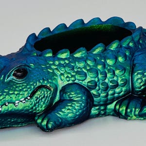 May include: A decorative alligator-shaped planter in shades of blue and green. The planter has a textured, scaly surface and an open top for plants. The alligator's eyes are black, and its mouth is slightly open, revealing small teeth.