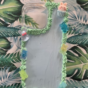 May include: A clear phone case with a light blue background and a green frosting border. The frosting is decorated with small blue and green gummy bears and a small pink bunny cupcake topper.