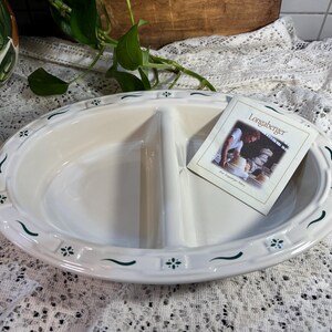 May include: Oval, divided serving dish with a white glaze and green floral accents. The dish has a decorative border and includes a small card with the name "Longaberger". The dish is designed for serving food.