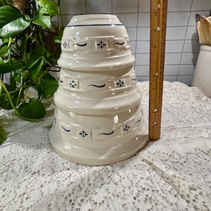 May include: A set of three stacked off-white ceramic mixing bowls with blue floral and geometric patterns. A wooden ruler placed vertically next to the bowls indicates a total height of approximately 12 inches for the stacked set.