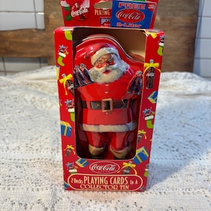 May include: A red Coca-Cola collector's tin featuring a Santa Claus figure. The tin box has a rectangular shape with a Santa Claus figure inside. The box also has the Coca-Cola logo and text that reads "2 Decks Playing Cards in a Collector Tin."