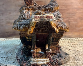 Peut inclure: Une miniature détaillée d'un cottage avec un toit de chaume et des fondations en pierre. L'enseigne indique "Bearly Able Construction Co." et "If you can dream it, we can build it." Le cottage est peint dans des tons bruns et beiges.