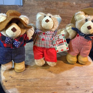 May include: Three plush teddy bears dressed in different outfits. One bear wears a brown hat, plaid shirt, denim overalls, and a bow tie. Another bear is in a plaid shirt and red shorts. The third bear has a straw hat and red and white striped overalls.
