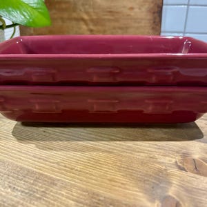 May include: Two rectangular, deep-dish ceramic baking pans in a solid dark red color. The exterior sides of the pans feature a raised, embossed basket-weave pattern. The dishes have a smooth, glossy finish and are stacked directly on top of each other.