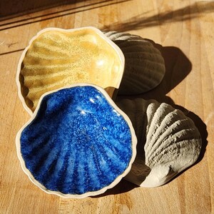 May include: A collection of four shell-shaped ceramic dishes. One dish features a vibrant blue glazed interior, another has a yellow glazed interior, and two are unglazed, matte off-white shells with detailed ridges and scalloped edges.