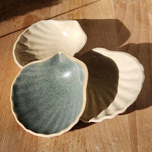 Puede incluir: Cuatro cuencos de cerámica con forma de concha marina en tonos blancos, verdes y marrones. Los cuencos están dispuestos sobre una superficie de madera, con sombras creadas por la luz. Los cuencos tienen un diseño texturizado y acanalado.