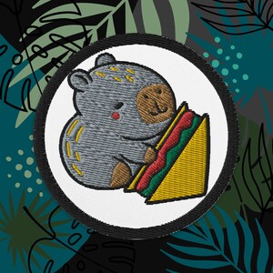 May include: A gray embroidered patch with a cartoon animal holding a yellow and green taco. The animal has brown ears and a brown nose.