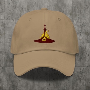 May include: A tan baseball cap with an embroidered design of a sword sticking into a skull with flames on either side. The skull and flames are yellow, and the sword is red.