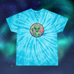 Può includere: Una t-shirt tie-dye con un alieno verde che indossa un segno di pace su uno sfondo colorato.