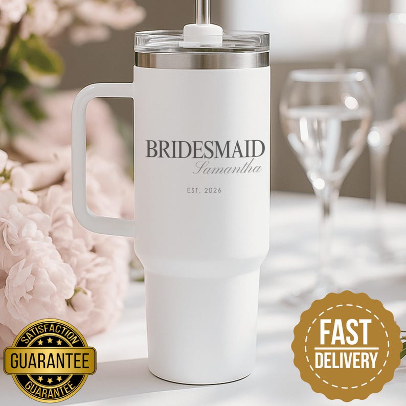 May include: A white stainless steel tumbler with a handle, a clear lid, and a straw. The front features grey text reading BRIDESMAID, Samantha, and EST. 2026. The design is clean and functional, with a tapered base and a secure lid mechanism.