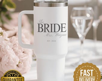 Può includere: Un bicchiere bianco in acciaio inossidabile con manico e coperchio trasparente dotato di cannuccia. La parte anteriore presenta la scritta The BRIDE, Mrs. Wayne e EST. 2026 in un carattere serif grigio scuro sul corpo cilindrico.