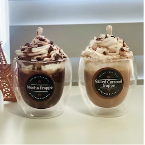 May include: Two frappe-shaped candles in clear glass cups. One is mocha, topped with chocolate chips and whipped cream. The other is salted caramel, with caramel pieces and whipped cream. Each has a label that reads "Natural Soy Wax" and the frappe flavor.
