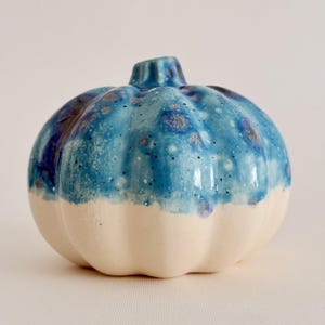 Op de afbeelding: Een keramische pompoensculptuur met een glanzend, gevlekt blauw glazuur op de bovenste helft met donkerblauwe en witte spikkels, overgaand in een gladde, matte, ongeglazuurde crèmekleurige basis met verticale ribbels en een kleine steel.