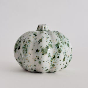 Op de afbeelding: Een keramieken pompoenfiguur met een glanzend witte glazuurlaag voorzien van een gespikkeld patroon met donkergroene, lichtgroene en kleine gele stippen. Het object heeft een ronde, geribbelde vorm met een kleine, textuurrijke steel.