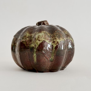 Op de afbeelding: Een keramische pompoensculptuur met een glanzende afwerking, voorzien van een diepbruine basiskleur en een lichtere saliegroene glazuurlaag die vanaf de bovenste steel naar beneden druipt, wat zorgt voor een textuurrijk uiterlijk.