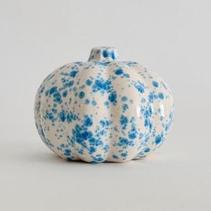 Pode incluir: Uma figura de abóbora em cerâmica com uma base branca brilhante e um padrão de manchas azul vibrante. O objeto tem uma forma arredondada e canelada com um pequeno caule distinto no topo e um acabamento liso e reflexivo.