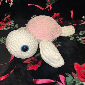 May include: A crocheted turtle toy with a pink shell and white body. The turtle has a blue eye and is lying on a black and pink floral fabric.