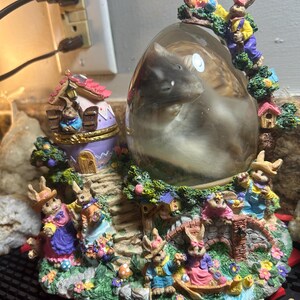 May include: A decorative Easter-themed snow globe featuring a sleeping animal inside a clear egg-shaped enclosure. The base is adorned with colorful bunny figurines, a bridge, and a miniature house. The scene is filled with vibrant colors and intricate details.