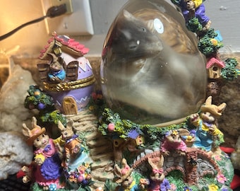 May include: A decorative Easter-themed snow globe featuring a sleeping animal inside a clear egg-shaped enclosure. The base is adorned with colorful bunny figurines, a bridge, and a miniature house. The scene is filled with vibrant colors and intricate details.