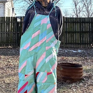 May include: A person wearing a green, pink, and blue patchwork overall with a black hooded sweatshirt and a brown beanie. The overall has a geometric pattern and is made of different fabrics.