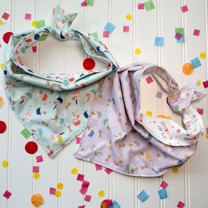 May include: Two dog bandanas with a birthday theme. One bandana is light blue with a cupcake and bone pattern. The other bandana is purple with a cupcake and bone pattern and the text "It's My Birthday!" in rainbow letters.