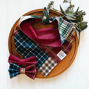 May include: A dog bandana and bow tie set in a variety of plaid patterns, including burgundy, green, and gold. The bandana has a snap closure. The bow tie is burgundy with a plaid center. The set is displayed on a wooden tray.