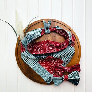 May include: A red, blue, and white bandana with a checkered pattern. The bandana is tied into a bow and is sitting on a wooden surface.