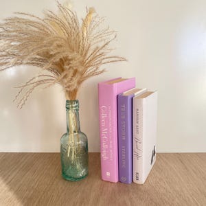May include: A still life arrangement with a green glass bottle vase holding dried pampas grass, alongside three hardcover books in pink, purple, and white. The books display titles and author names on their spines, creating a decorative display.