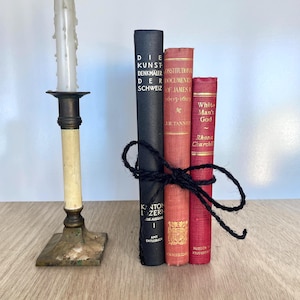 Puede incluir: Un candelabro de latón antiguo con una vela color crema se encuentra junto a una pila de tres libros antiguos atados con hilo negro. Los libros son en tonos rojos y negros.